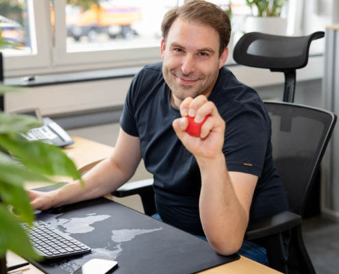 Am Arbeitsplatz, Mitarbeiter schaut lächelnd in die Kamera und hat einen roten Stressball in der Hand, Teamfotos für Unternehmenswebsite