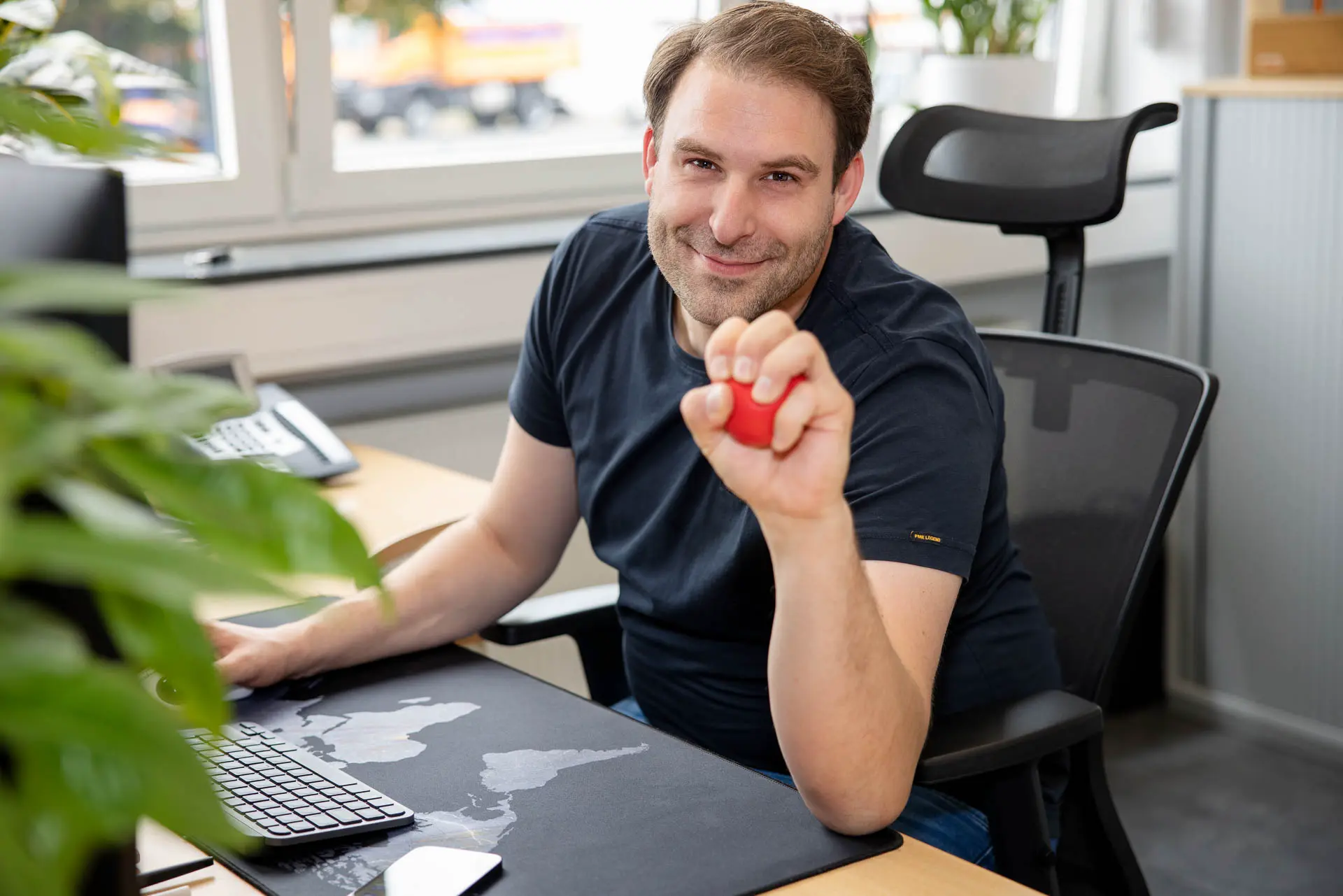 Am Arbeitsplatz, Mitarbeiter schaut lächelnd in die Kamera und hat einen roten Stressball in der Hand, Teamfotos für Unternehmenswebsite