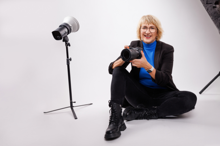 Eine professionelle Fotografin, lächelnd und entspannt sitzend, hält eine Spiegelreflexkamera in einem gut ausgestatteten Fotostudio. Sie trägt eine schwarze Hose, Stiefel, ein blaues Oberteil und eine schwarze Jacke, was ihr ein stilvolles Erscheinungsbild verleiht. Im Hintergrund ist ein Teil der Studioausrüstung zu sehen, darunter ein Beleuchtungsstativ. Das Bild strahlt Professionalität und Leidenschaft für die Fotografie aus.