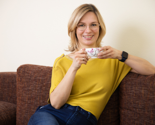 Personal Branding Fotoshooting mit Anne Buckler - systemische Coachin - Koblenz-Gülls - Sitzend mit Kaffeetasse, sie schaut freundlich in die Kamera, gelbes Top und Jeans