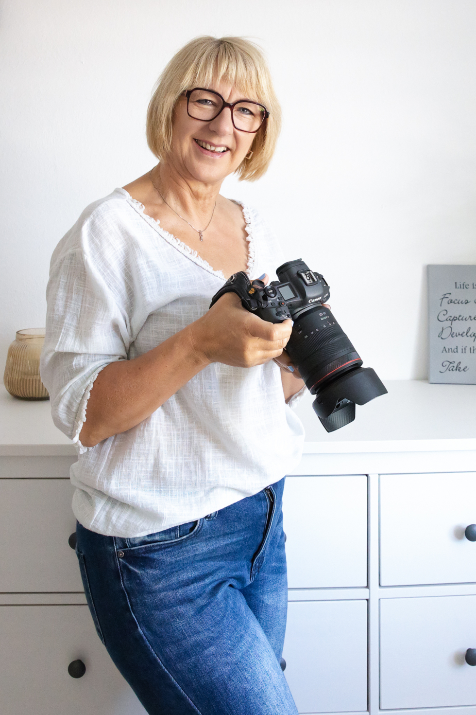 Foto mit prof. Kamera - Fotografin mit der Kamera schaut freundlich in die Kamera. Sie trägt eine weiße Bluse, rote Brille, blaue Jeans und hat blonde Haare. Hinter ihr ist eine weiße Kommode und weiße Wand.