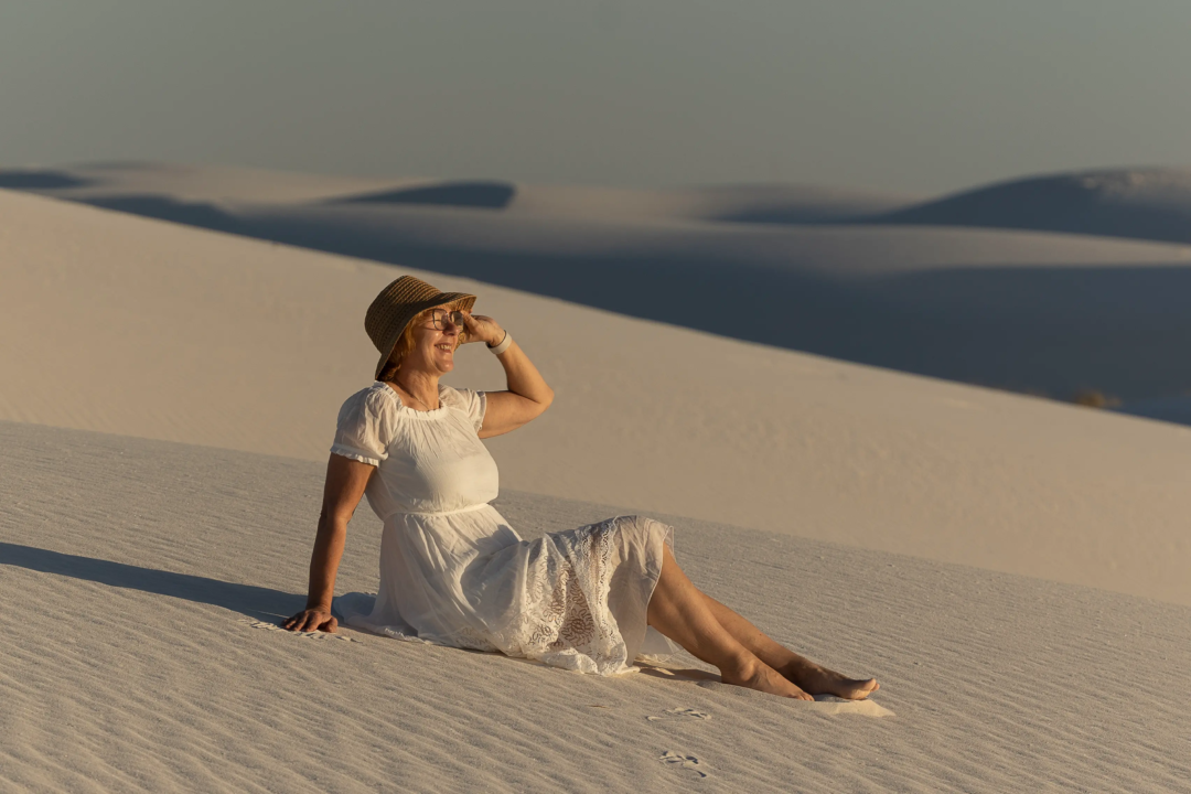 Zeig uns dein Lieblingsfoto von dir und erzähle die Geschichte dahinter. Karina Schuh im weißen Kleid und Strohhut sitzt auf einer weißen Sanddüne im White Sands Nationalpark, genießt den Sonnenuntergang und schaut in die Ferne.