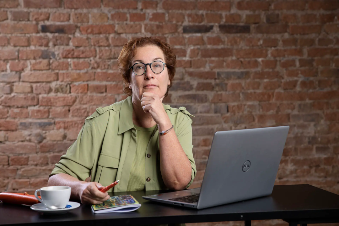 Autorin Carla Capellmann am Laptop. Sie hat eine Hand am Kinn und schaut nach oben und überlegt. In der Hand hält sie einen Kugelschreiber. Eine Tasse Kaffee steht mit auf dem Schreibtisch. Sie trägt eine grüne Bluse. Authentische Markenfotografie für die Website.