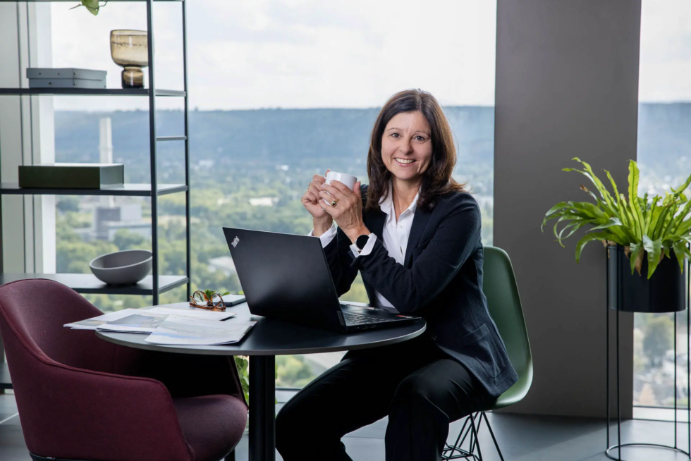 Immobilienmaklerin mit der Kaffeetasse in der Hand, vor ihr der Laptop, in der 27. Etage am Kanzlerplatz in Bonn und schaut freundlich in die Kamera - Personal Branding Fotografie für Corporate Influencer im Arbeitskontext