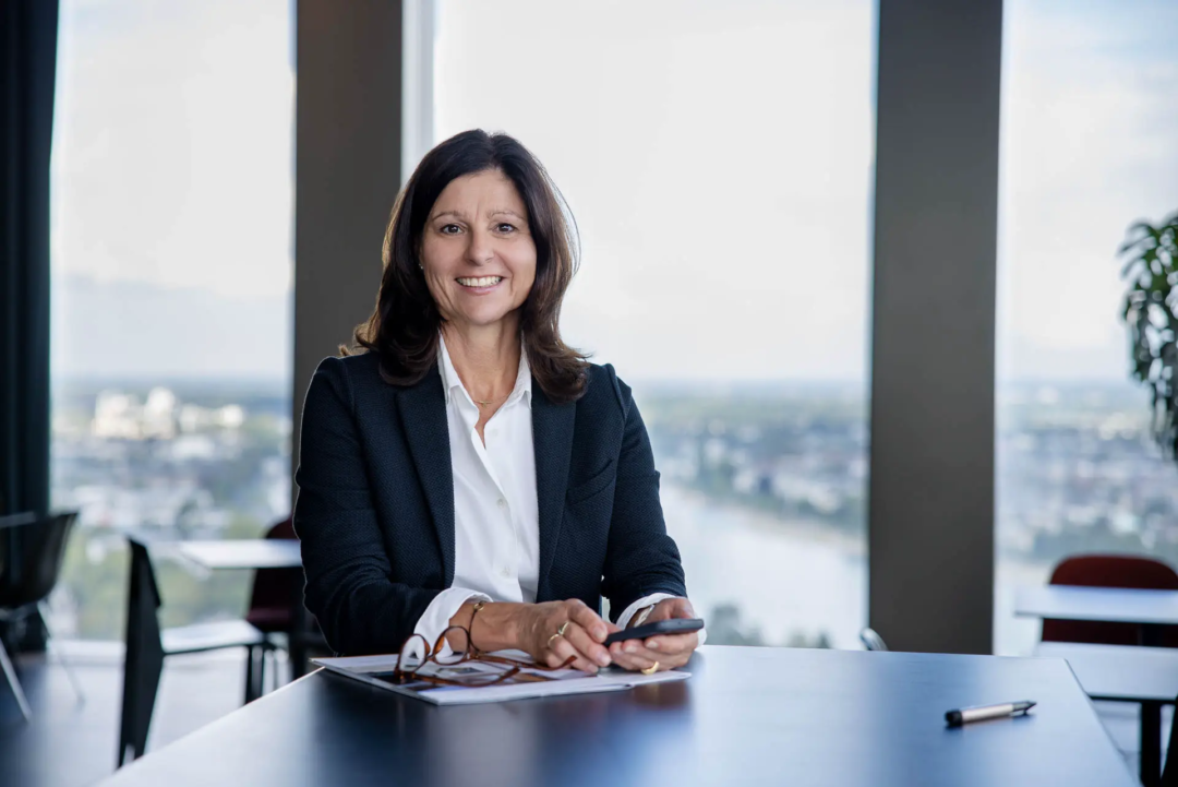 Personal Branding Fotos für die Über-Mich-Seite von Claudia Ewenz, Immobilienmaklerin in Bonn, authentisches Portrait der Maklerin mit dem Smartphone in der Hand. Sie steht am Tisch und hinter ihr sieht man den Rhein und die Stadt Bonn.