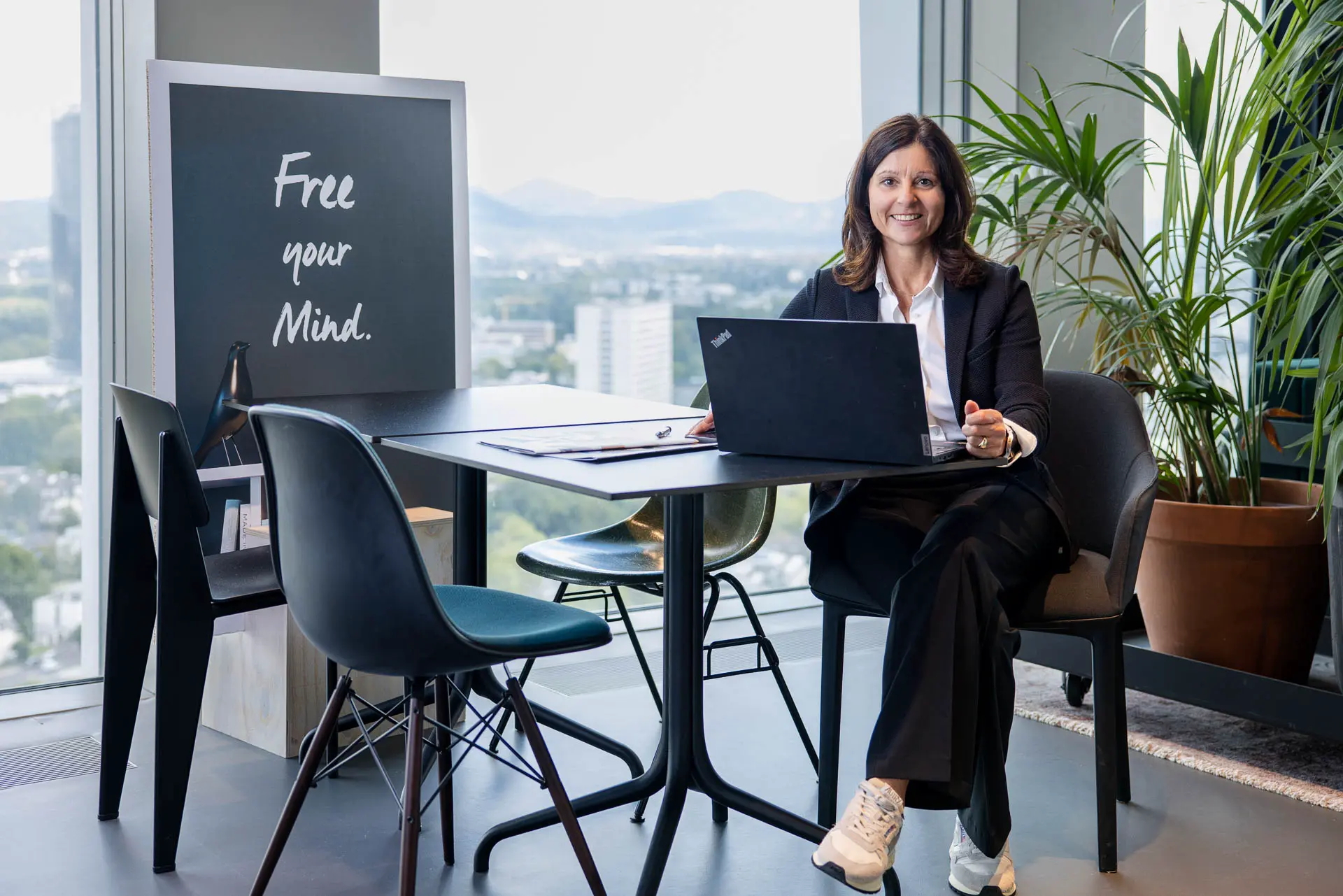 Immobilienmaklerin sitzt am Tisch, vor ihr der Laptop, in der 27. Etage am Kanzlerplatz in Bonn und schaut freundlich in die Kamera