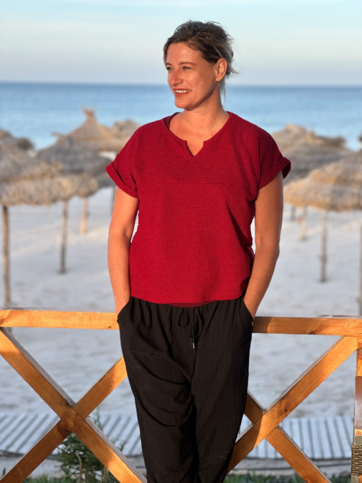 Antje Backwinkel, Buchautorin, steht auf einer Holzveranda am Strand, mit einer leichten Brise im Haar. Sie trägt ein bordeauxrotes Oberteil und eine schwarze Hose. Im Hintergrund sind Schilfhütten und der Strand zu sehen. Virtuelles Fotoshooting am Strand von Djerba.