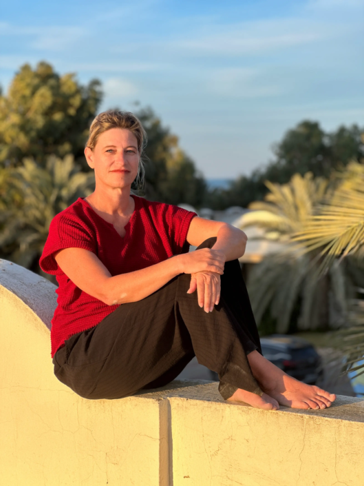 Antje Backwinkel, Buchautorin, sitzt entspannt auf einer Mauer im Abendlicht, trägt ein bordeauxrotes Oberteil und schwarze Hose. Hinter ihr sind Palmen und eine friedliche Umgebung sichtbar. Virtuelles Fotoshooting am Strand von Djerba.