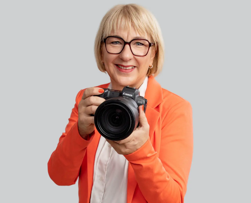 Fotografin Karina Schuh - Expertin für Employer Branding Fotografie - Hier mit der Kamera in der Hand. Sie trägt ein orangefarbenes Jacket , eine weiße Bluse und eine Jeans sowie eine Brille. Sie hat kurze blonde Haare und schaut freundlich in die Kamera