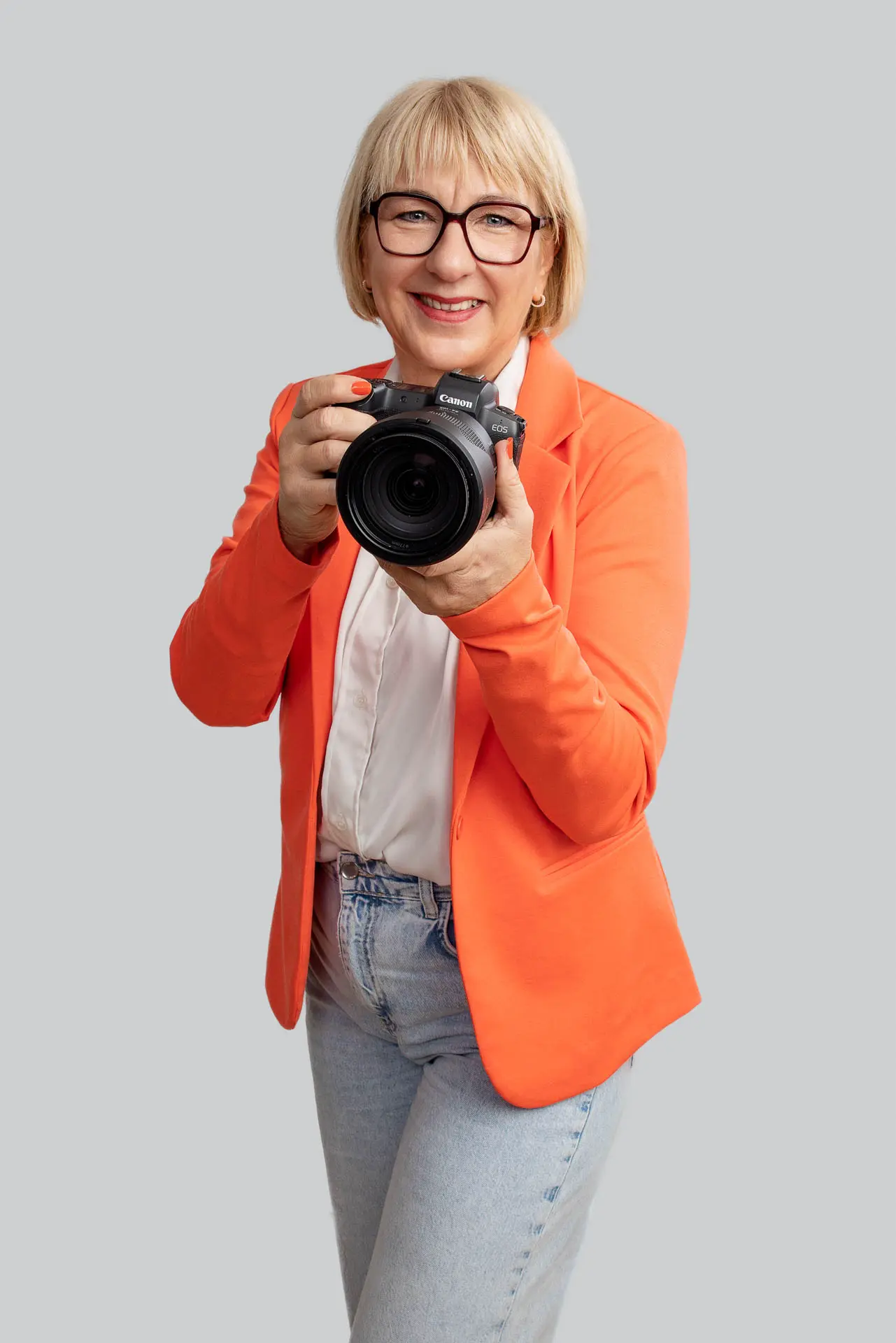 Fotografin Karina Schuh - Expertin für Employer Branding Fotografie - Hier mit der Kamera in der Hand. Sie trägt ein orangefarbenes Jacket , eine weiße Bluse und eine Jeans sowie eine Brille. Sie hat kurze blonde Haare und schaut freundlich in die Kamera