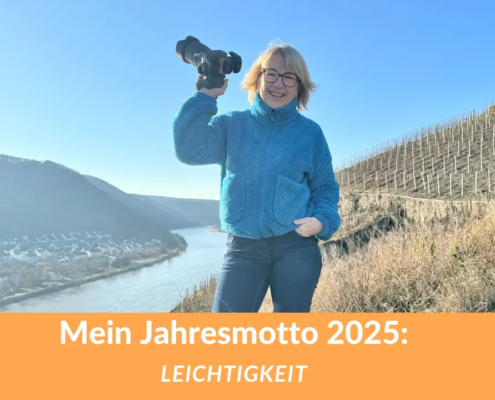 Karina Schuh, Fotografin, in einer entspannten Pose mit Kamera in der Hand, lächelnd vor einer malerischen Landschaft hoch über der Mosel. Text: Mein Jahresmotto 2025 – Leichtigkeit.