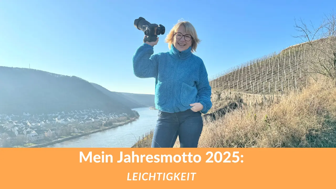 Karina Schuh, Fotografin, in einer entspannten Pose mit Kamera in der Hand, lächelnd vor einer malerischen Landschaft hoch über der Mosel. Text: Mein Jahresmotto 2025 – Leichtigkeit.