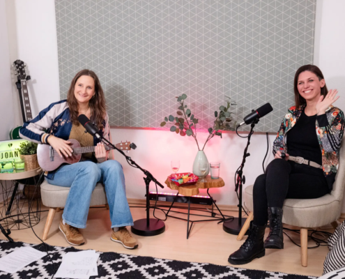 Joanna Gypser spielt auf ihrer Ukulele und lächelt, während Natalie Juwan neben ihr sitzt und freundlich in die Kamera winkt.