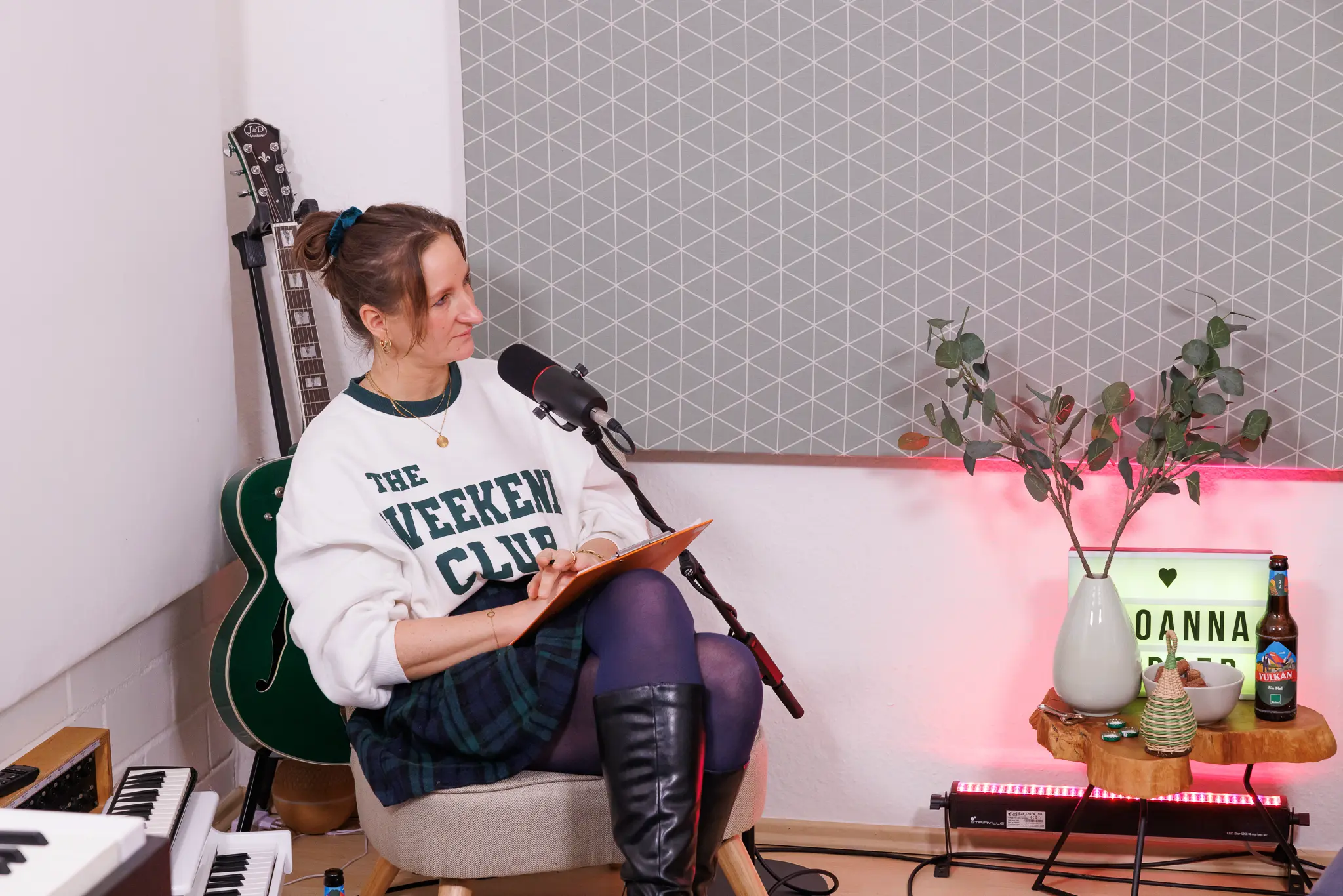 Joanna Gypser hält Notizen in der Hand und hört aufmerksam zu, während sie vor einem Mikrofon sitzt. Im Hintergrund sind eine grüne Gitarre und Dekorationen zu sehen.