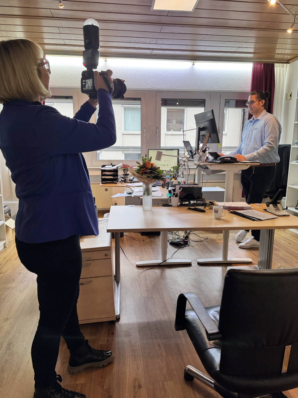 Eine Fotografin in blauer Jacke fotografiert einen Mann an seinem höhenverstellbaren Schreibtisch in einem modernen Büro. Der Arbeitsplatz ist mit Monitoren, Dokumenten und einem Blumenstrauß ausgestattet, während Tageslicht durch große Fenster einfällt.