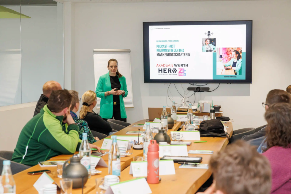 Ein Workshop-Szenario mit einer Frau in einem grünen Blazer, die vor einer Gruppe von Menschen steht und eine Präsentation auf einem großen Bildschirm hält. 