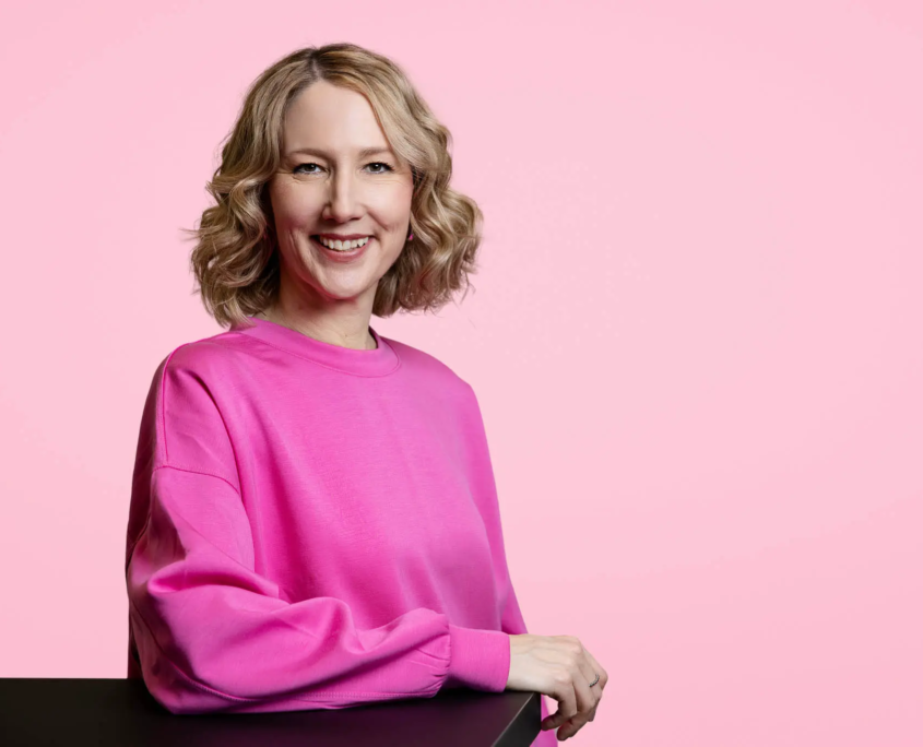 corine brötz in pinkfarbenem outfit vor pinkem hintergrund, freundlich lächelnd, blickt in die kamera, arm auf dem tisch – professionelles business portrait
