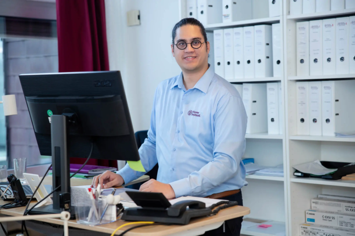 Mitarbeiter mit Brille arbeitet an einem großen Monitor im Büro, umgeben von Aktenordnern und Dokumenten.