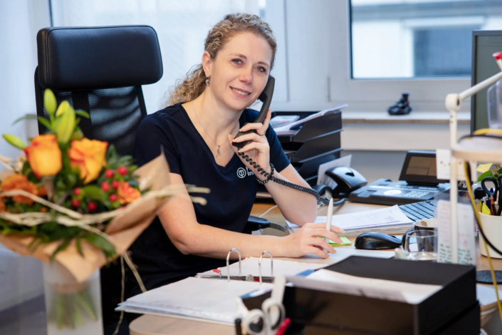 Mitarbeiterin mit Locken telefoniert an ihrem Schreibtisch, umgeben von Arbeitsunterlagen und einem Blumenstrauß.