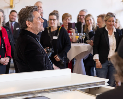 Thomas Anders lauscht einer musikalischen Darbietung am weißen Flügel, während die Gäste gespannt zuhören.