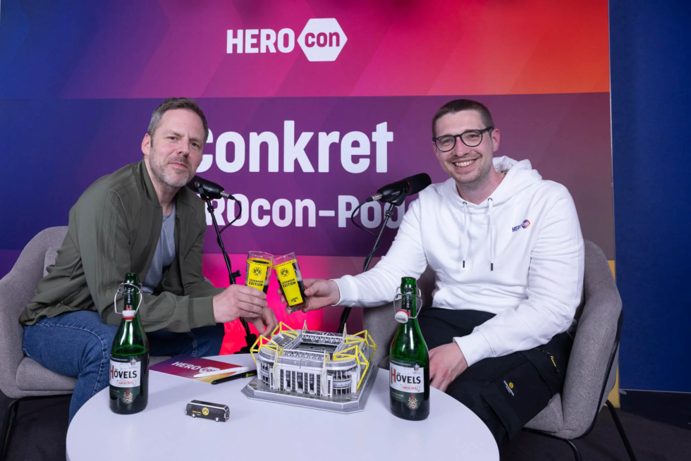 Robert Bittig und Niklas Palm von HERO Software beim HEROCON-Podcast 2025 mit BVB-Glas und Stadionmodell im Signal Iduna Park.