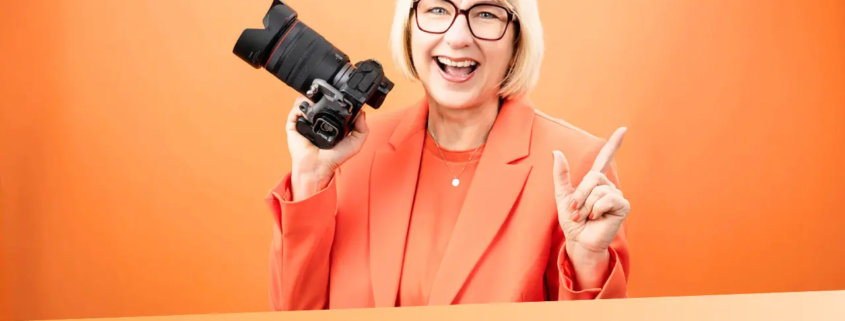 Karina Schuh mit Kamera vor orangefarbenem Hintergrund – Personal Branding Fotografie Markenbild